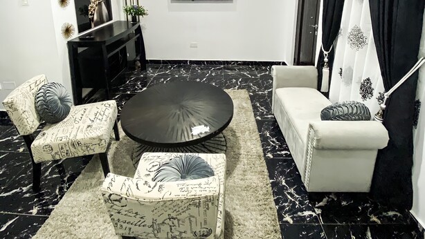 A modern living room with a round black coffee table, two patterned armchairs, a white sofa, and a flat-screen TV mounted on the wall. - Exquisite 1-bedroom Condo (Lagos)