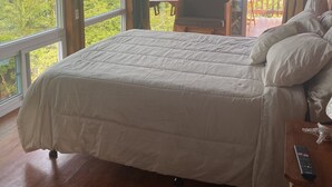 Makuuhuoneessa on suuri sänky, puulattia, parveke, jossa on pöytä ja tuolit, sekä näkymä merelle ja vehreään maisemaan.