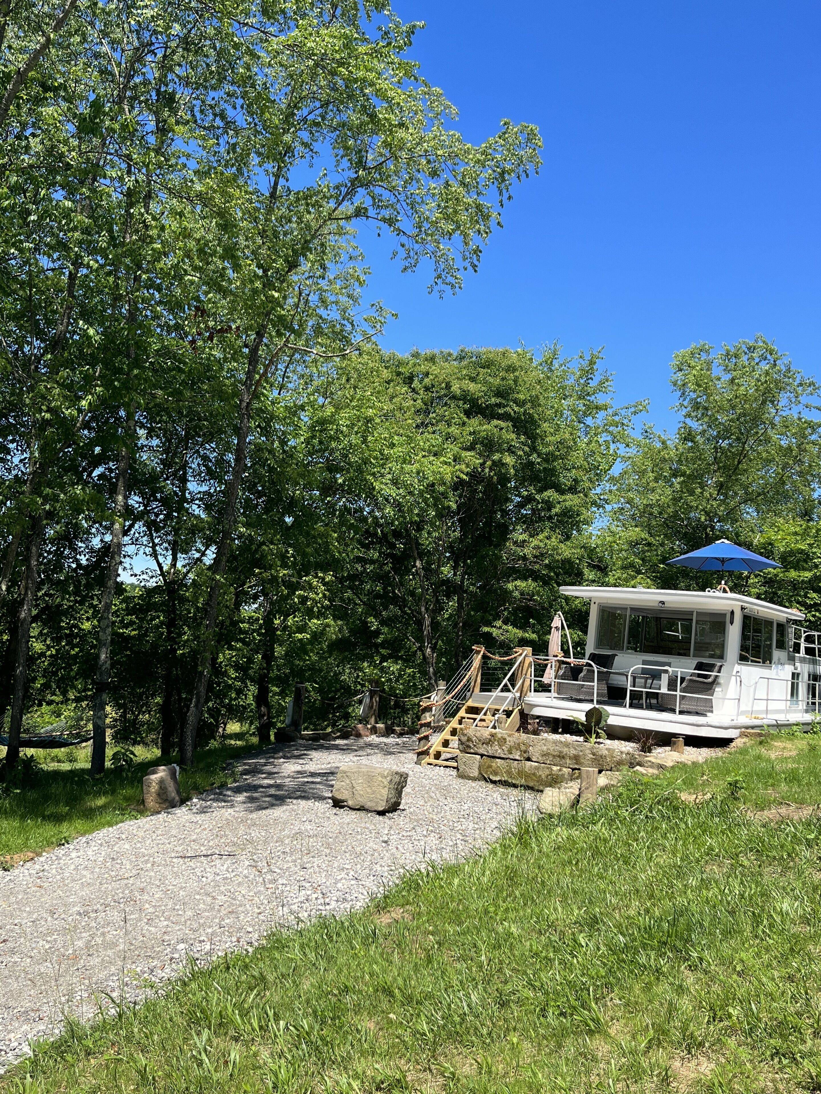 A boat docked near a gravel path surrounded by trees, with a blue umbrella and outdoor seating.