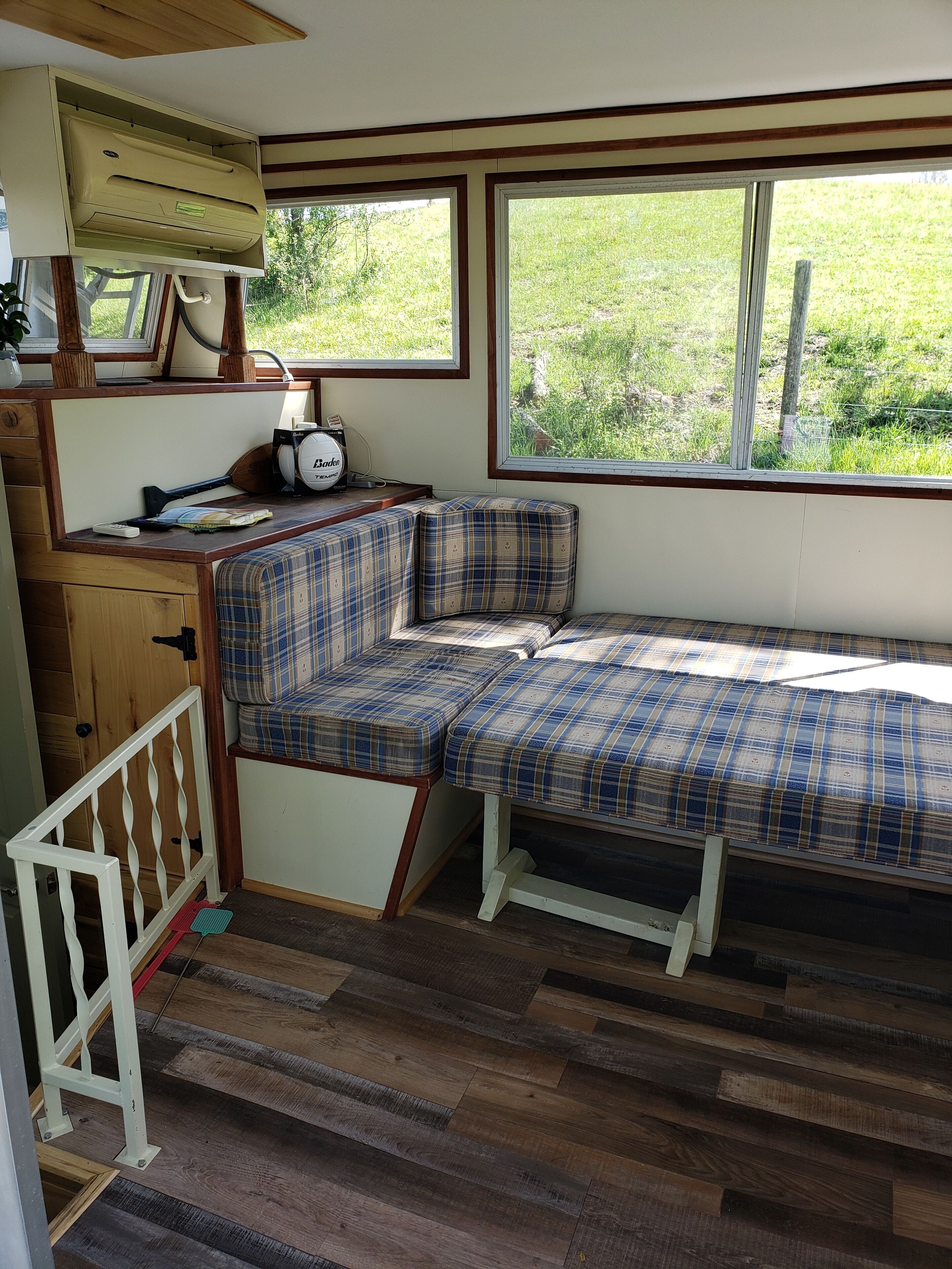 A cozy RV interior with a plaid bench, a small kitchenette, and a window offering a view of greenery.