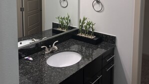 A bathroom with a black granite countertop, a white sink, and a mirror above it. There are two small potted plants on the countertop. - A lovely and well spaced one bedroom with a pool view (Houston)