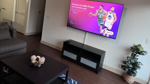 A living room with a flat-screen TV mounted on the wall displaying an NBA Finals advertisement. - A lovely and well spaced one bedroom with a pool view (Houston)