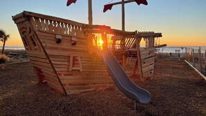 A wooden pirate ship play structure with a slide, set against a sunset background. - ~Gorgeous~ Ocean View King Suite in Cabana Row~ (Myrtle Beach)