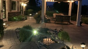 A well-lit patio area with a stone pathway leading to a seating area under a pergola, surrounded by landscaped greenery and a small water feature. - A Taste of Tuscany -Just 5 Minutes to Golf and Wine Tasting  (Fallbrook)