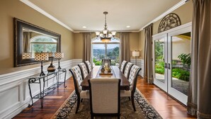 A dining room with a long table, six chairs, a chandelier, and a view through a glass door. - A Taste of Tuscany -Just 5 Minutes to Golf and Wine Tasting  (Fallbrook)