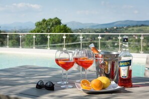 Una mesa con tres vasos de bebida roja, una botella de Aperol y limones en un balcón con piscina y montañas al fondo.