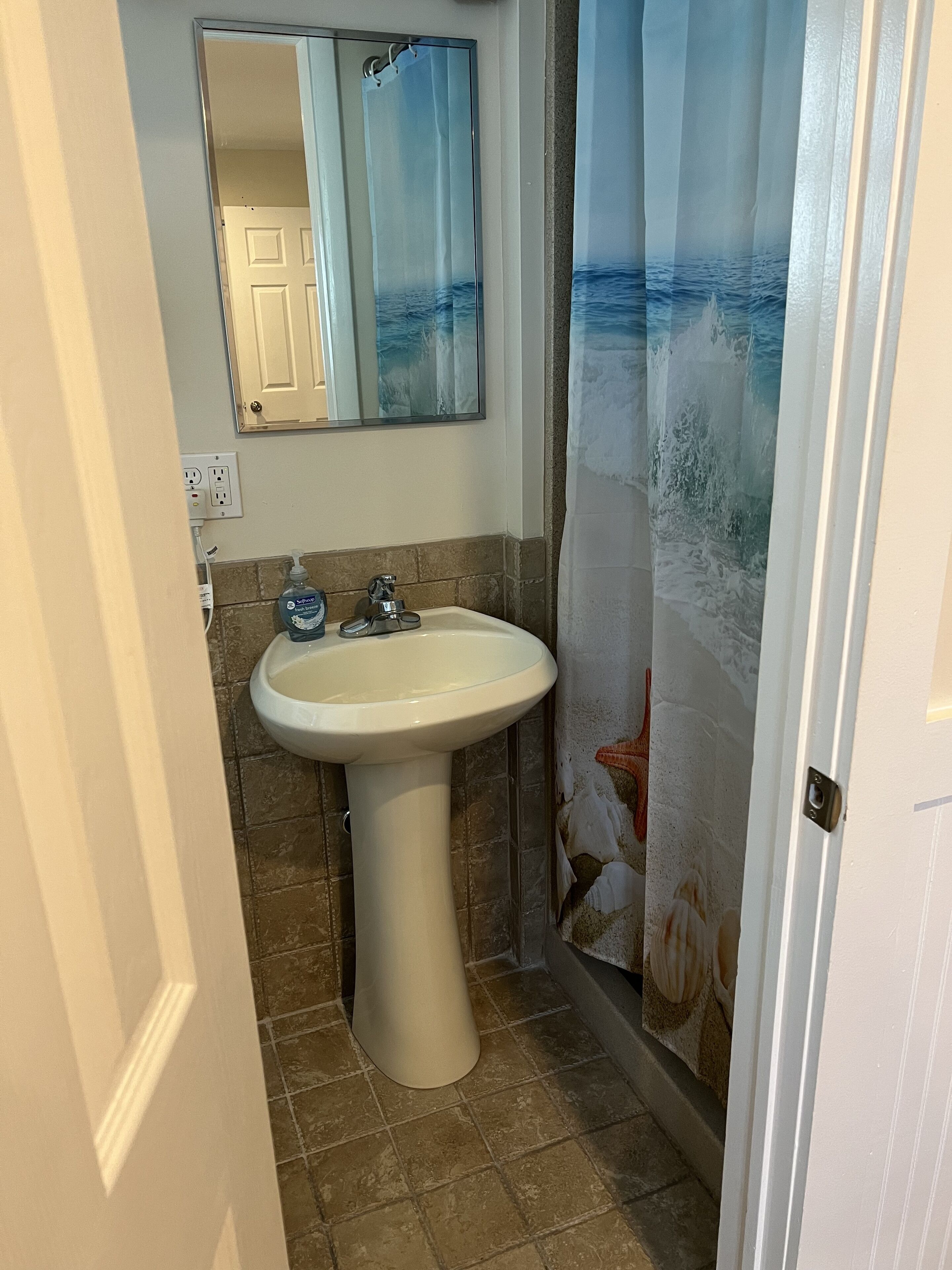 Un baño con lavamanos pedestal, un espejo encima y cortina de ducha con escena de playa.