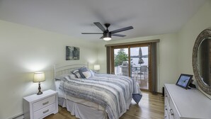 A bedroom with a bed, bedside table, lamp, ceiling fan, and a view of a balcony with outdoor furniture.