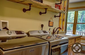 Ein Waschbereich mit zwei Maytag Waschmaschinen und Trocknern, Holzregalen mit Haken, einem Fenster mit Blick auf Bäume und einem Schild mit der Aufschrift 'live'.