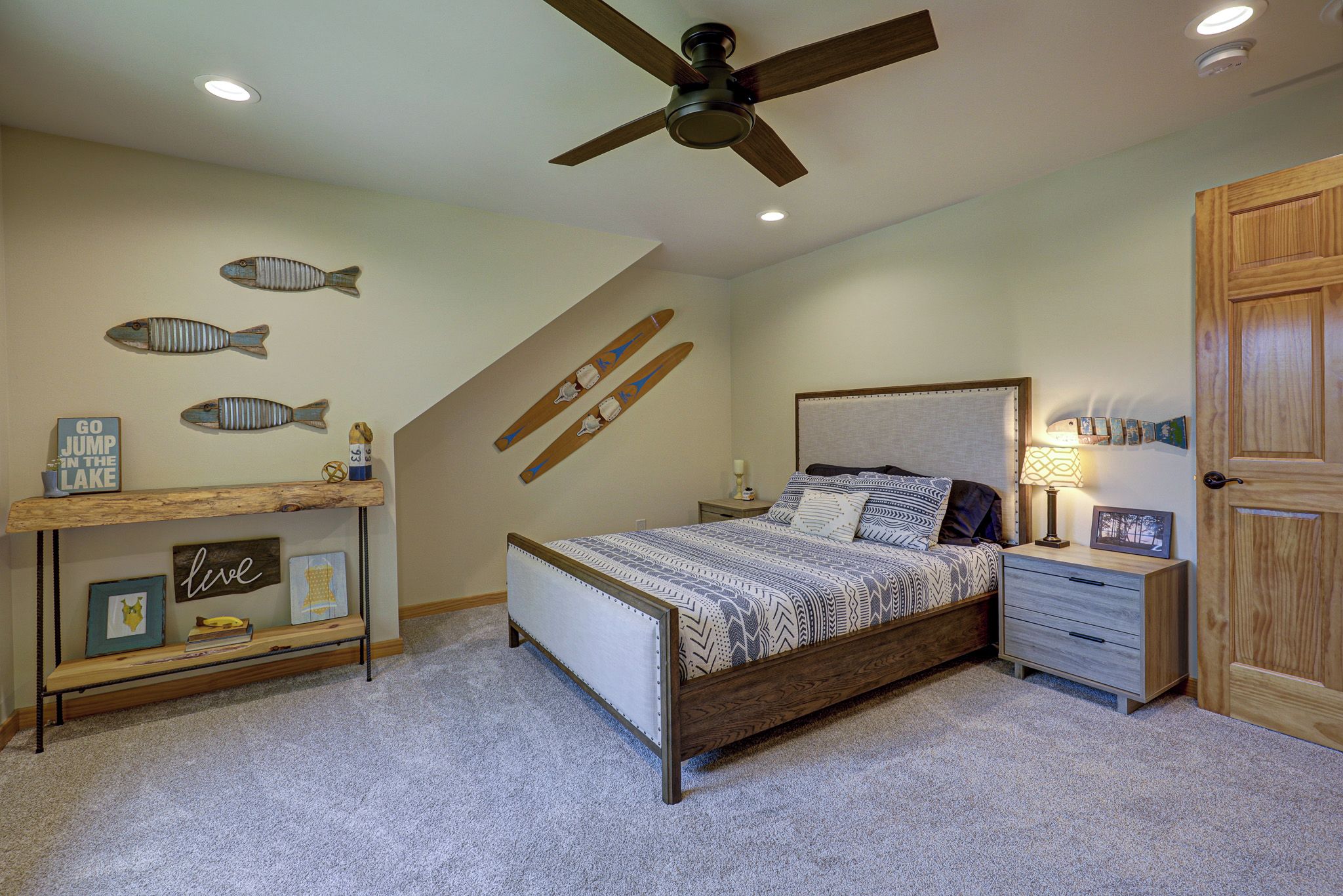 A bedroom with a bed, a nightstand, a desk, a ceiling fan, and wall decorations including fish and skis.
