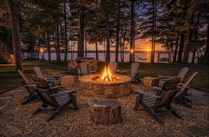 Un foyer circulaire, entouré de fauteuils Adirondack, installé sur une surface de gravier, avec vue sur un lac et un coucher de soleil en arrière-plan.