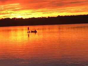 Un atardecer sobre una masa de agua tranquila con siluetas de una persona de pie y otra en un bote.