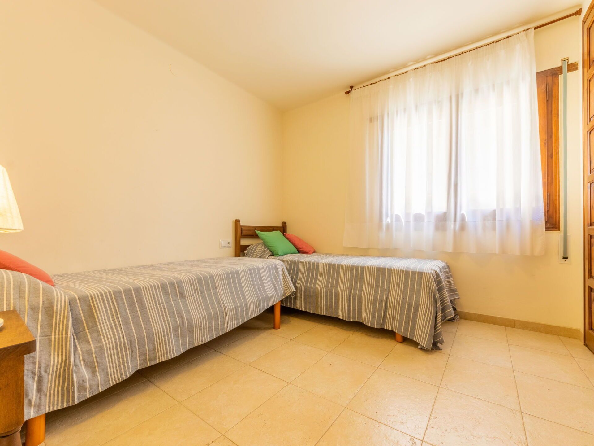 A small, bright room with two single beds, a wooden nightstand, a lamp, and a window with sheer curtains.