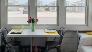 Uma área de refeições com uma mesa posta para dois, um vaso com flores e vista para casas através de grandes janelas.