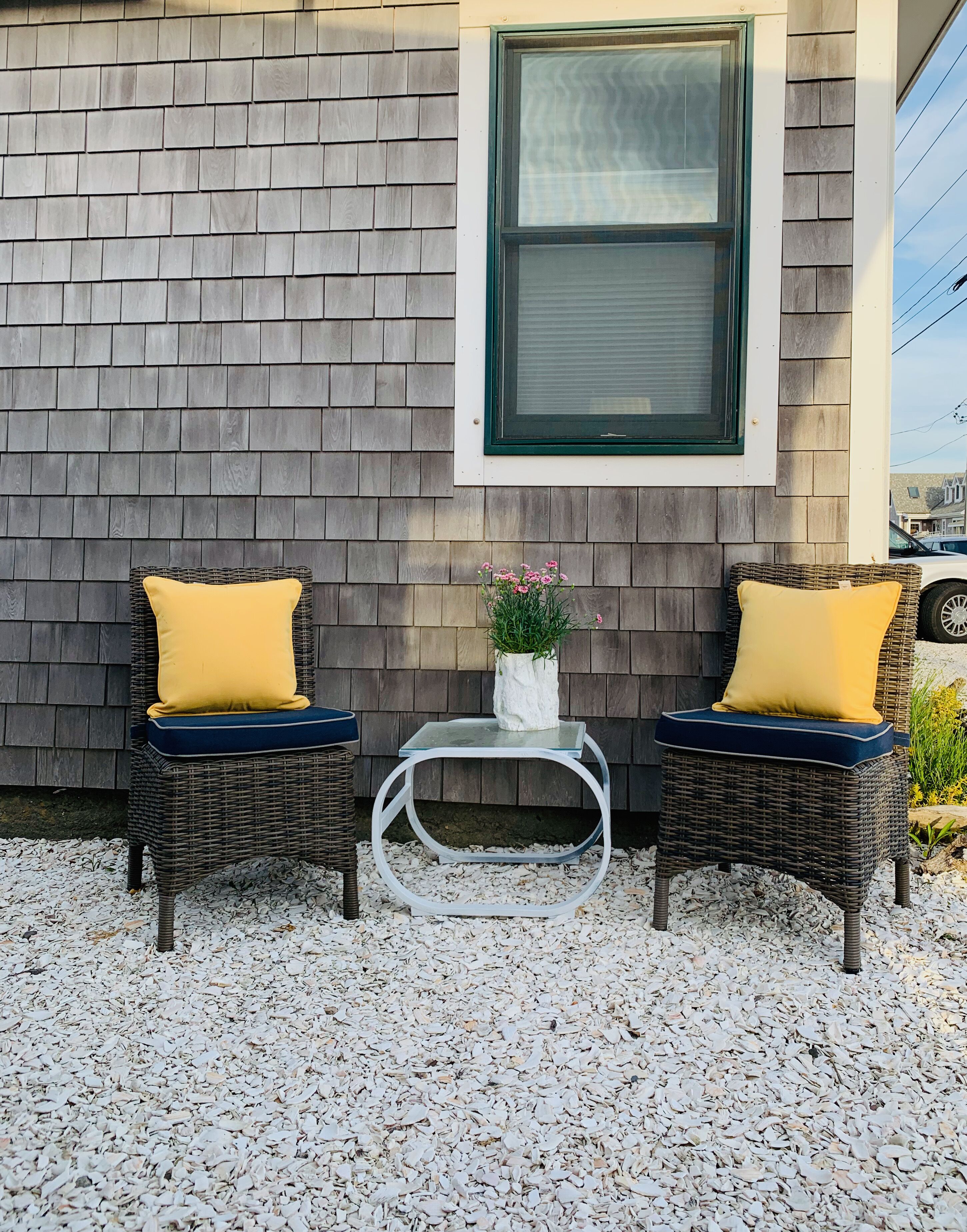 Two wicker chairs with yellow cushions and a small table with a vase of flowers between them, set on a gravel surface against a wooden wall with a window above.