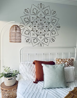 Ein Schlafzimmer mit einem weißen Bett, einem dekorativen Wandmuster, einem gewebten Korb mit einer Pflanze und einer hängenden Leuchte.
