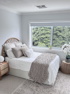 A bedroom with a large window, a bed with white bedding, a bedside table with a lamp, and a woven side table.