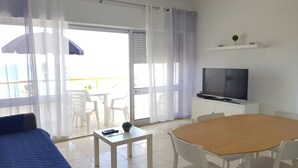 A hotel room with a bed, a TV, a dining table with chairs, a small table with a remote control, and a balcony with chairs and a table. - T1 Praiamar 4E A C FR MAR Garagem (Loulé)