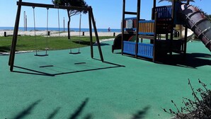A playground with swings and a slide, set against a beach backdrop. - T1 Avenida Mar 8 200m Praia Wi-fi 4 Pessoas (Loulé)