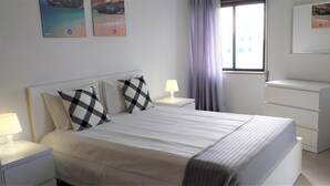 A neatly arranged bedroom with a bed, bedside tables, a dresser, and a window with curtains. - T1 Avenida Mar 8 200m Praia Wi-fi 4 Pessoas (Loulé)