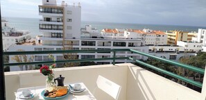A balcony with a table set for breakfast, overlooking a coastal cityscape with modern buildings and a view of the sea. - T1 Avenida Mar 8 200m Praia Wi-fi 4 Pessoas (Loulé)