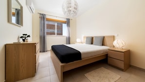 A bedroom with a bed, bedside tables, a dresser, a potted plant, and a window with curtains. - T1 Varandas Forte Piscina Ar Cond Garagem (Loulé)