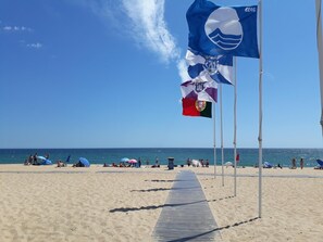 A beach with a wooden walkway leading to the sea, flags, and people relaxing. - T1 Varandas Forte Piscina Ar Cond Garagem (Loulé)