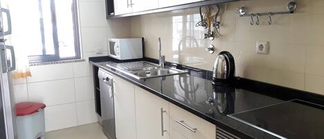 A kitchen with white cabinets, a black countertop, a microwave, a kettle, a sink, and a dishwasher.
