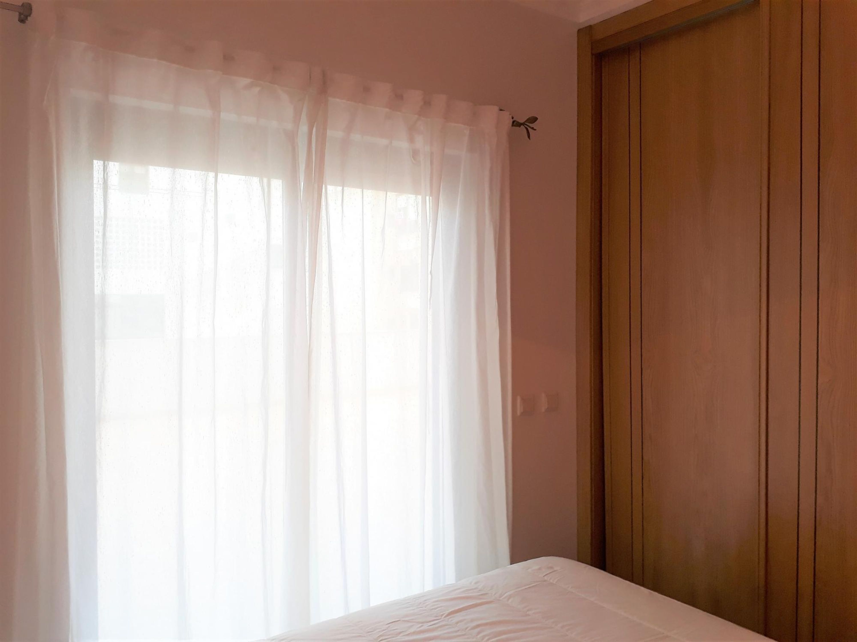 A bedroom with a wooden door, a white bed, and sheer curtains.