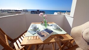 Ein Holztisch mit einem Buch, zwei Tassen und einer Blumenvase auf einem Balkon mit Blick aufs Meer.