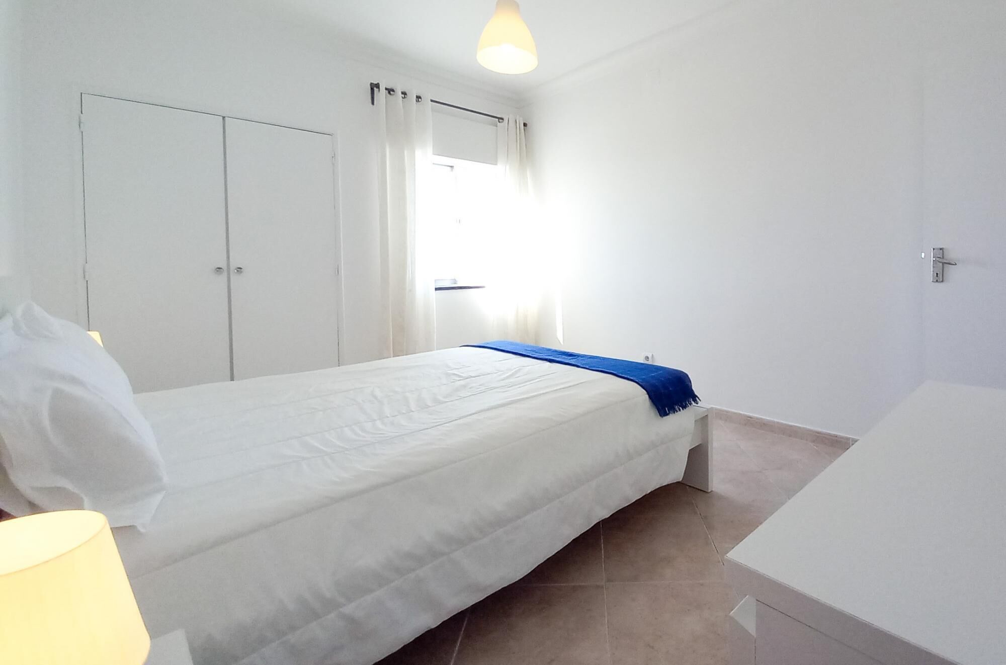 A bedroom with a bed, a wardrobe, a window with curtains, and a bathroom with a sink and mirror.