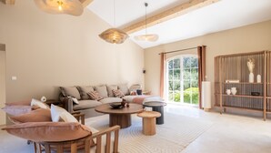 A spacious living room with a sofa, coffee table, and chairs, wooden beams on the ceiling, and a large window with curtains.