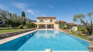 A large swimming pool with a stone border, a house with a tiled roof, and a well-maintained garden with trees and shrubs.