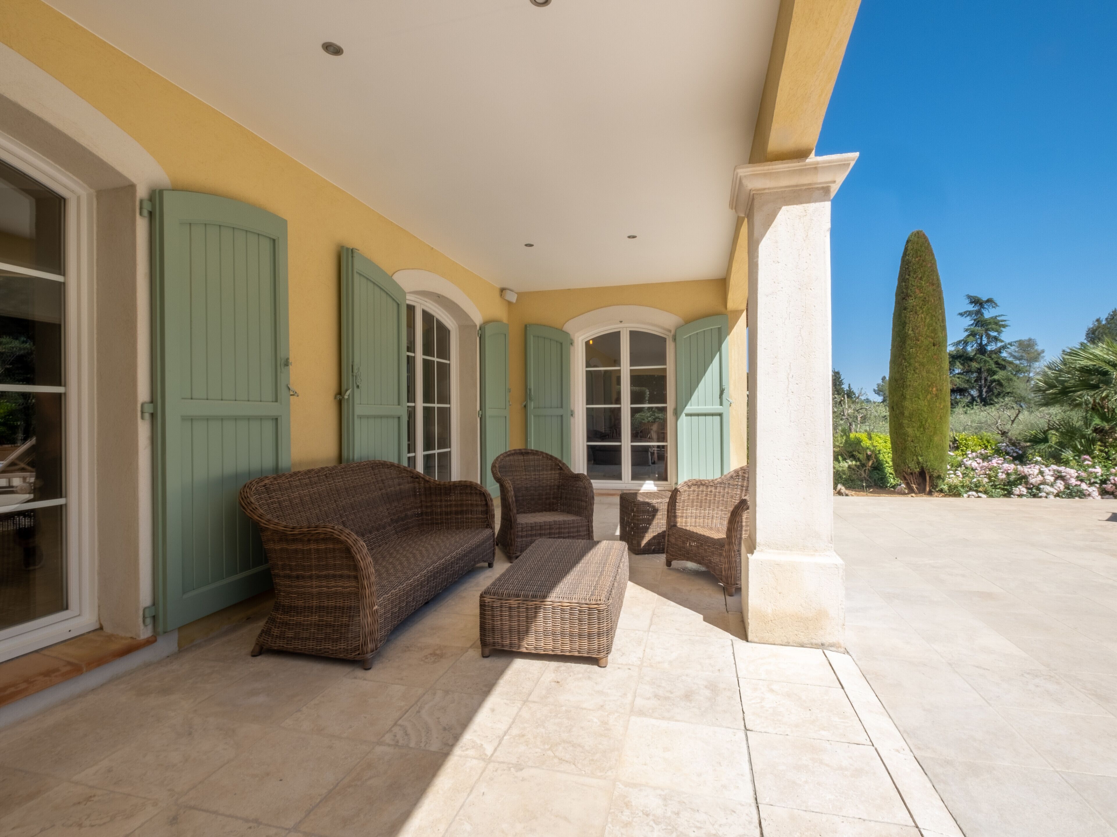 A covered patio area with wicker furniture, a tiled floor, and a view of a garden with a tall tree.