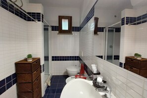 Un bagno con piastrelle bianche, un lavandino bianco, uno specchio, una piccola finestra e uno spazio doccia.