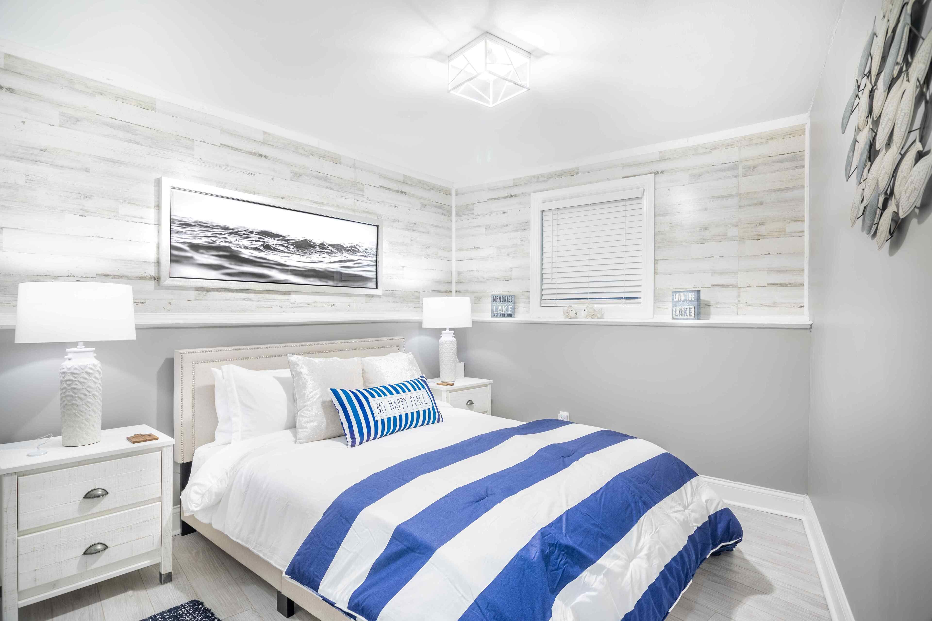 Een slaapkamer met een bed, nachtkastjes, een lamp, een lijst met een afbeelding en een raam met jaloezieën.