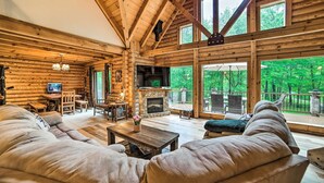 A log cabin interior with a stone fireplace, wooden furniture, and large windows overlooking a forest. - Classy Home w/ Hot Tub: 2 Mi to Lake Access! (Jackson)