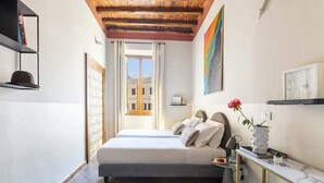 Une chambre à coucher comprenant un lit, une table de chevet, une commode, une fenêtre avec des rideaux, un tableau au mur et une vue sur les bâtiments à l’extérieur.