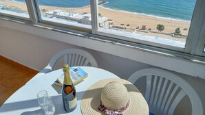 A table with a bottle of champagne, a book, and a straw hat by a window overlooking a beach.