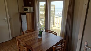 Une table à manger en bois avec quatre chaises, une orchidée en pot, un four à micro-ondes posé sur un meuble, et une vue sur la mer depuis un balcon équipé de stores en bois.