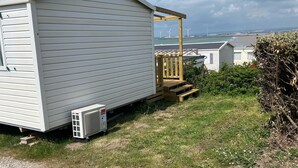 Une maison mobile dotée d’un porche couvert et d’une petite terrasse, située sur une pelouse avec un chemin de gravier devant.