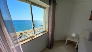 A bedroom with a large window overlooking a beach, a bed, a bedside table with a lamp, and a curtain. - T1 Torre Azul 11 Vista MAR Fabuloso P R DO SOL WI- (Loulé)