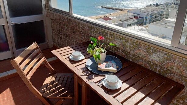 A balcony with a wooden table, two white cups, a book, and a potted plant. - T1 Torre Azul 11 Vista MAR Fabuloso P R DO SOL WI- (Loulé)