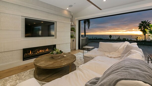 A modern living room with a fireplace, a large TV, and a view of the sunset over the ocean.