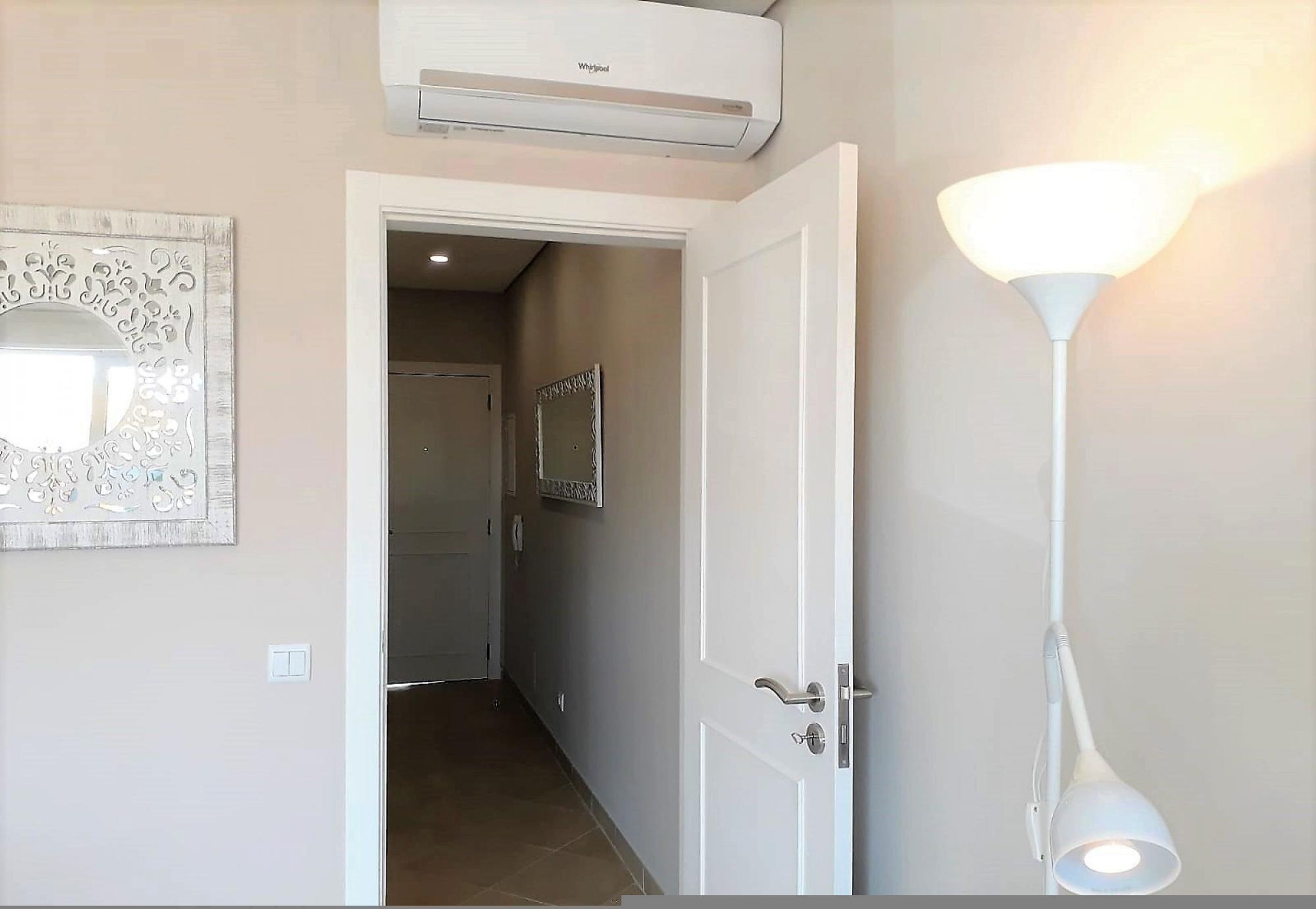 A hallway with a white door, a wall-mounted air conditioner, and a wall-mounted light fixture.