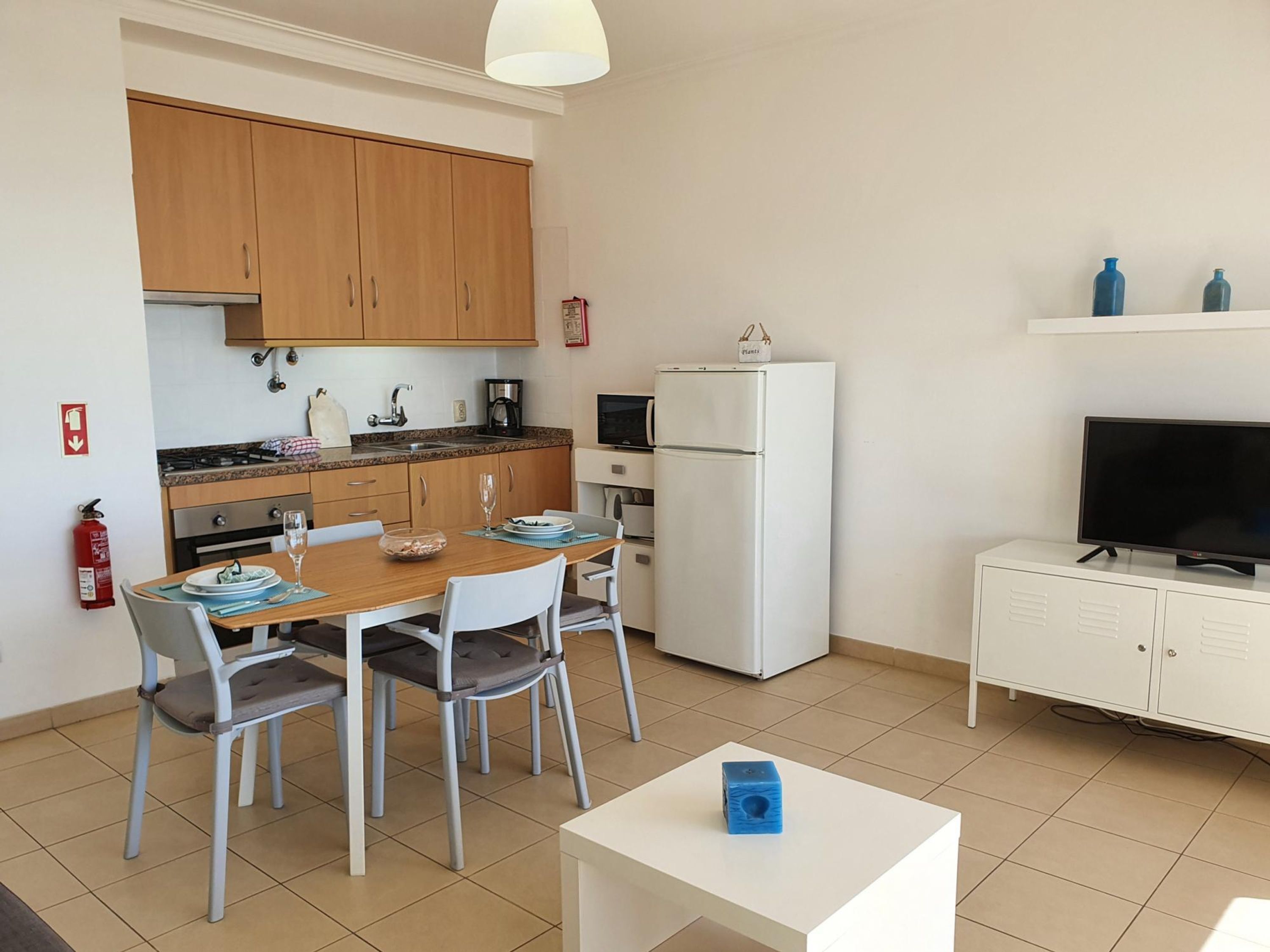 A compact kitchen with wooden cabinets, a white refrigerator, a microwave, and a coffee maker. There is a dining table with chairs and a small white cabinet with a television.