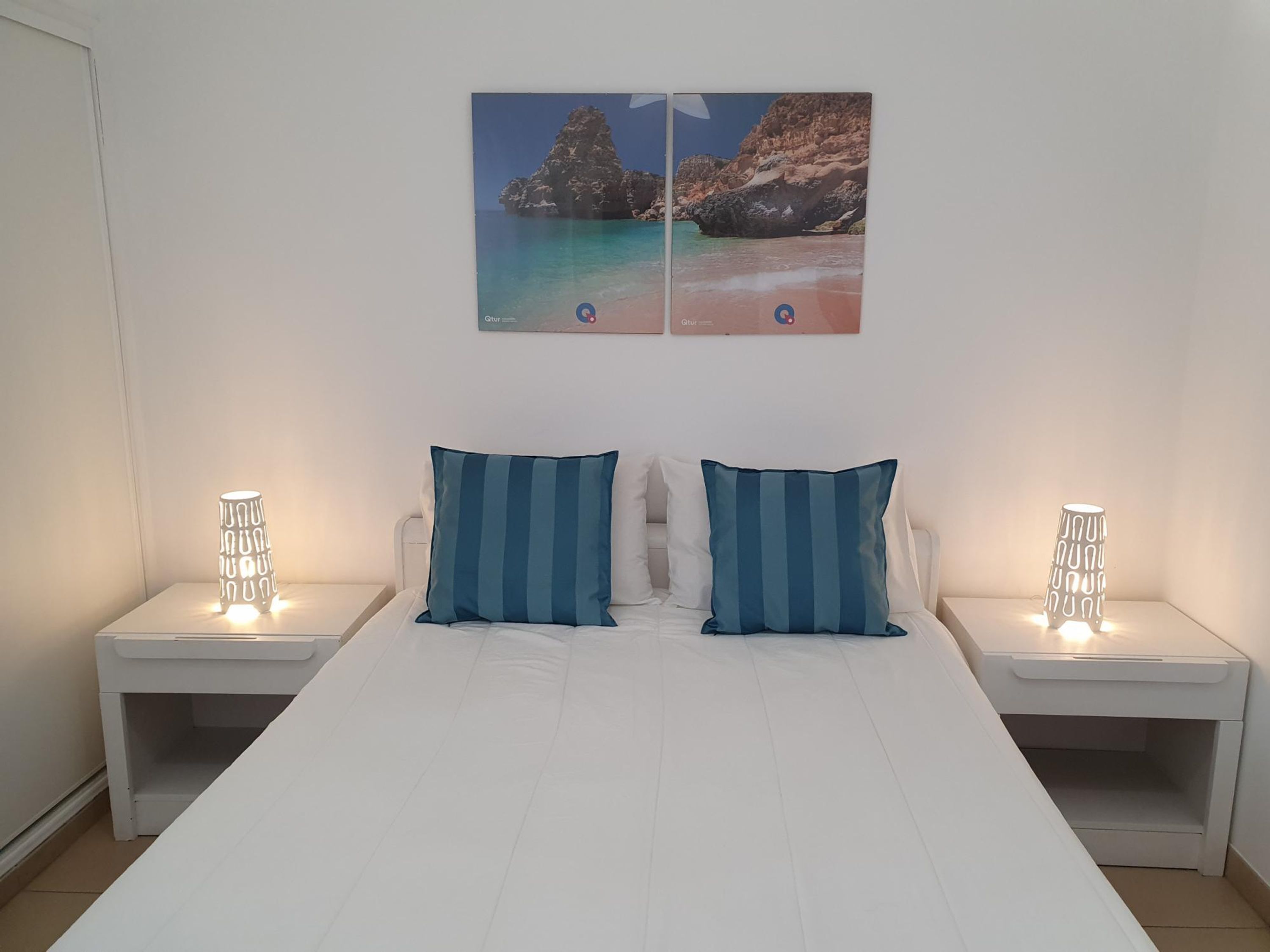 A bedroom with a bed, two nightstands, two blue-striped pillows, and a wall-mounted picture of a beach scene.