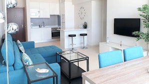 A modern living room with a blue sofa, a glass coffee table, a TV, and a kitchen in the background. - T1 Conforto E Vista MAR A C 80M Praia-16e Wifi (Loulé)