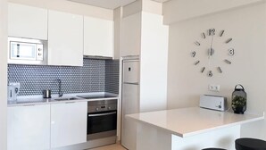 A modern kitchen with white cabinetry, a black and white backsplash, a built-in oven, a microwave, a refrigerator, a clock, and two black bar stools. - T1 Conforto E Vista MAR A C 80M Praia-16e Wifi (Loulé)
