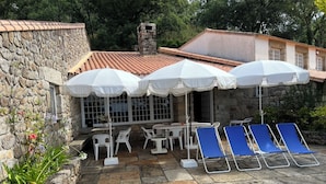 Een gebouw van steen met een pannendak, terras met witte en blauwe stoelen en witte parasols.